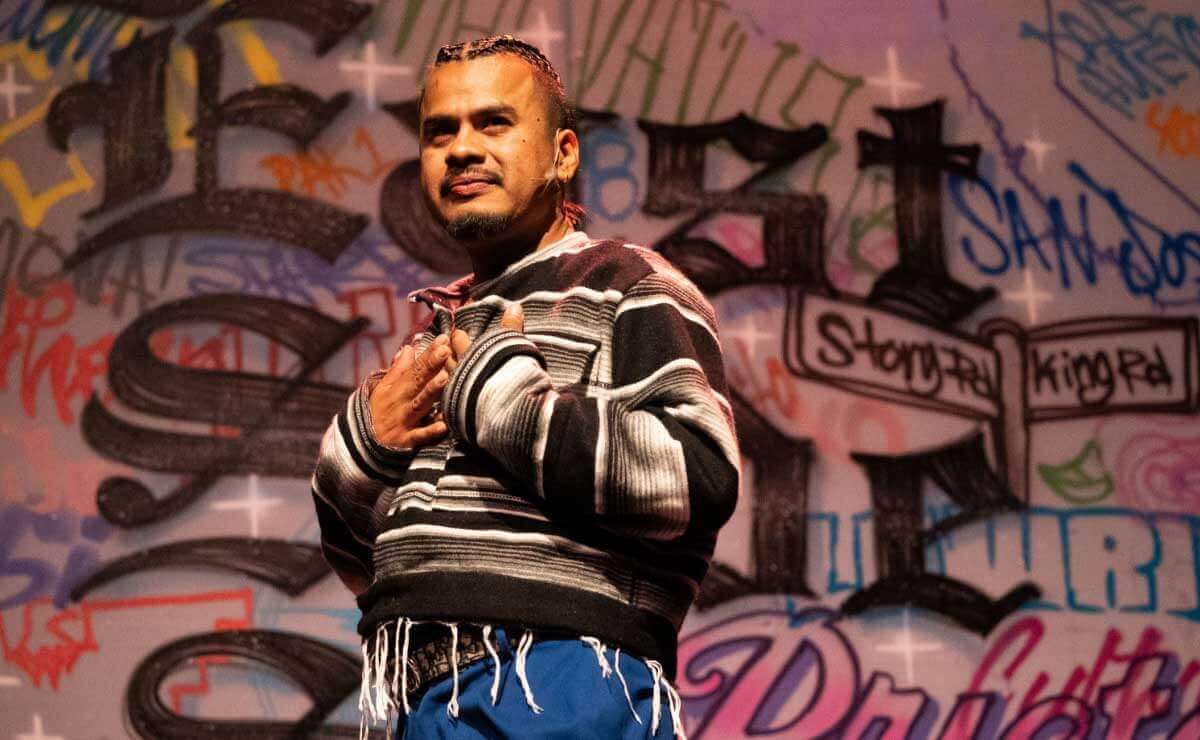 Close up of a medium-brown skinned man staring off into the distance, wearing a striped black and grey jacket, with blue pants, and in the background, a wall of graffiti.