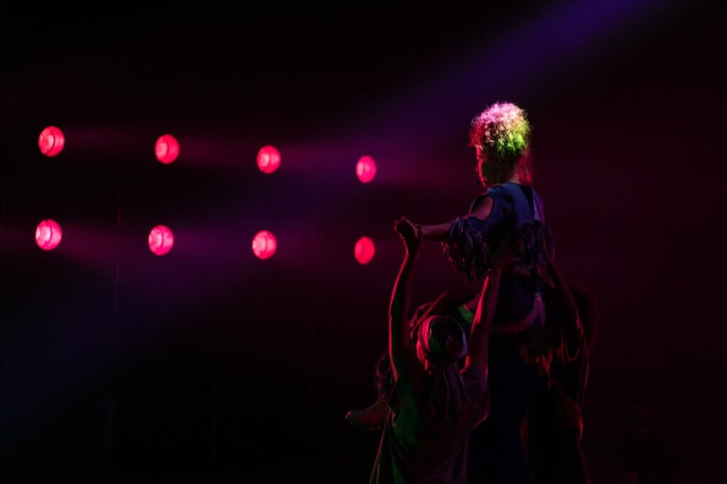 A light-skinned curly-haired haired femme in denim fringe is hoisted up onto the shoulders of another Black femme by two other Black femme performers -- one in a du-rag, one with her hair out in a soft afro. A row of pink lights shine on their faces, and they are backlit in an otherwise dark scene.