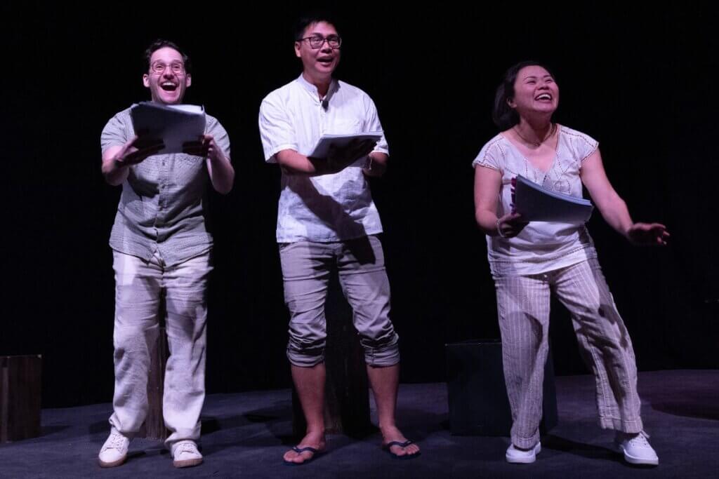 A trio of actors with scripts in hand perform in a reading of MIX-MIX - a white man with glasses, a medium light-skinned Filipino man with glasses, and a petite medium-skinned Filipino woman stare up to the sky, with wide joyful smiles on their faces. They wear pants and shirts of whites, tans, and light browns.