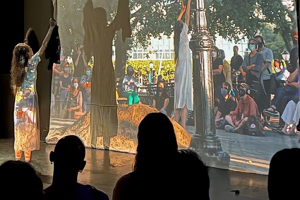 A light-skinned woman dancing towards a screen displaying a projected video of the woman’s live performance recorded earlier. She has both of her arms in the air and is carrying black cloth in both hands. She is wearing all white and is standing solo on the stage. The project's video displays her dancing similarly at Battery Park City.