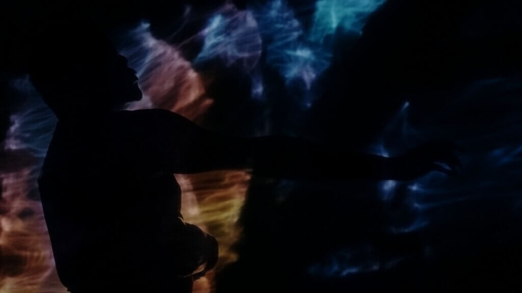 jaamil olawale kosoko's profile is in silhouette is set against a vibrant background of swirling, multicolored lights. An arm extends gracefully, creating a sense of movement and fluidity. The light, wispy patterns in the background blend shades of blue, purple, and orange, adding a dramatic and ethereal atmosphere to the scene.
