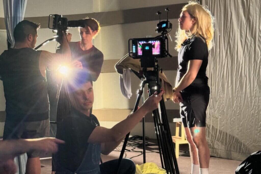 An image of four artists working with two cameras on tripods to create live camera feeds for a workshop of Happy Fall: A Queer Stunt Spectacular. On the left side of the photo, a medium-skinned gender-fluid artist holds the camera while a female light-skinned artist helps with the tripod. A second group of artists includes a medium-skinned male artist wearing overalls manning a tripod filming a male medium-skinned artist who is standing wearing a long blond female wig.