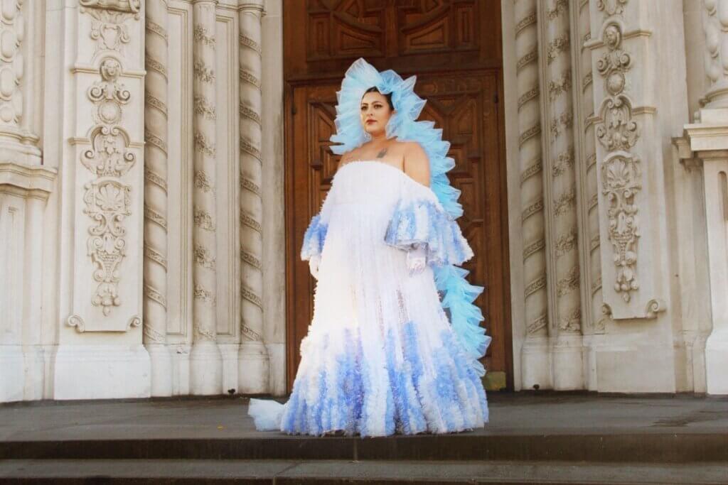 A Medium-skinned Latine woman stands in front of ornate cathedral doors and archway with hands at her sides looking outward. She is in a white gown with sky blue sleeves and gown bottom with a ruffle head piece that looks like a full body length halo.