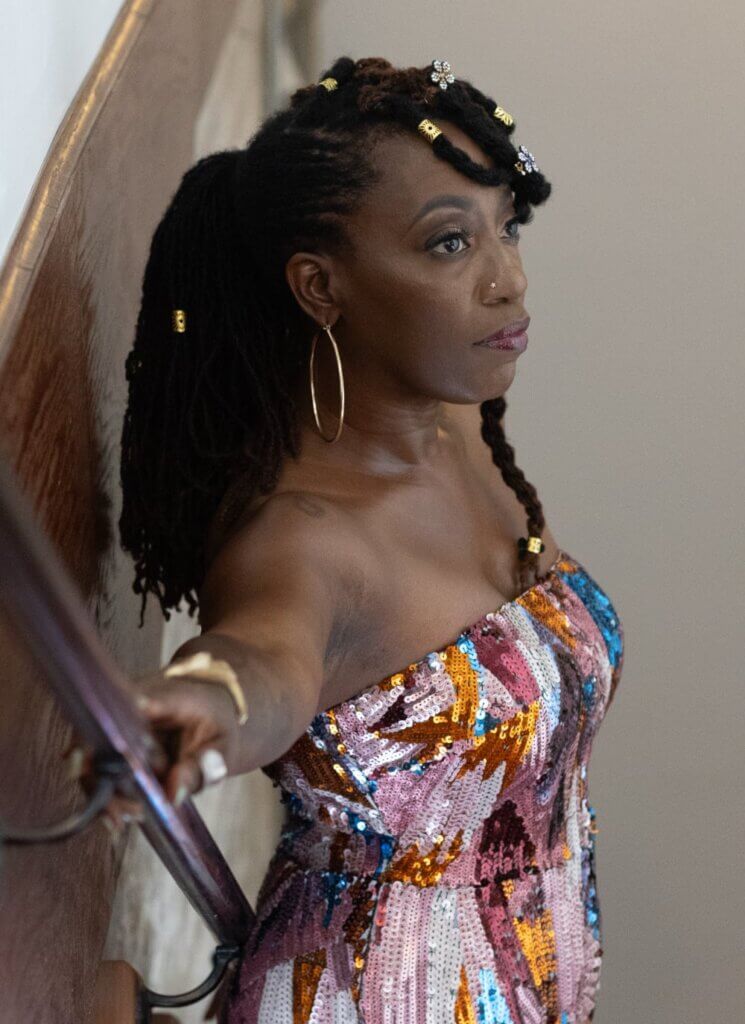 Erica Falls This is an image of a medium-dark-skinned Black woman standing in a stairwell with her back to the wall, looking away from the viewer with her arm extended on the railing. Her loc'd hair is in a ponytail. She is wearing a strapless, multi-colored sequin jumpsuit.