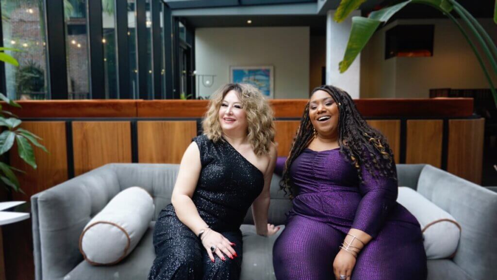 Two women sit on a modern-style gray velvet sofa in an upscale lounge. A wall of windows illuminates them from the left. There is green foliage on either side of the frame. On the left, Lubana Al Quntar has light skin and medium blonde shoulder-length wavy hair, wearing a sparkly black dress that is sleeveless and off the shoulder on the left side. On the right, Andrea “Vocab” Sanderson has medium-dark skin and long black hair in twists with gold highlights, wearing a sparkly royal purple jumpsuit. Both women are laughing, radiating a confident and light-hearted energy.