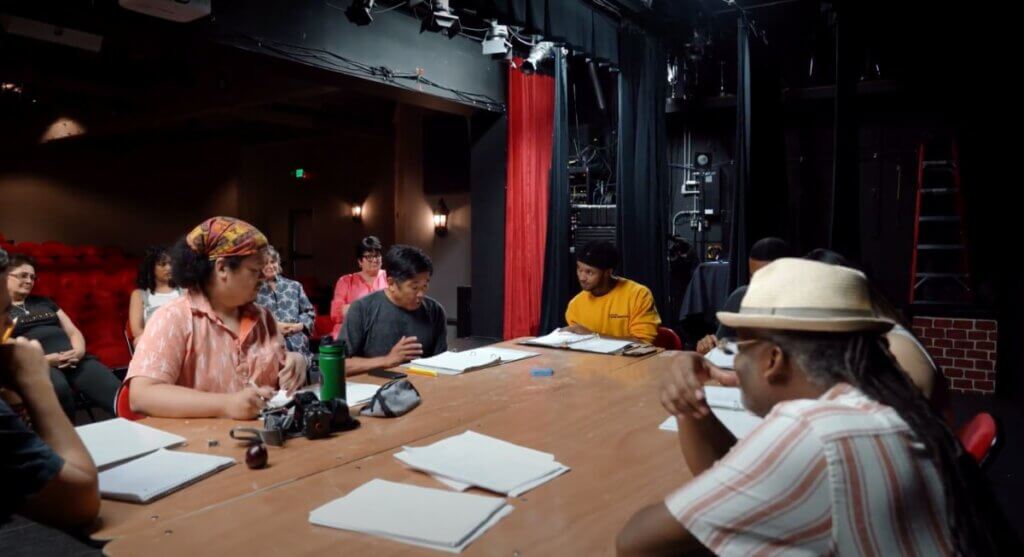 A table of Black and Japanese performers, a playwright, and director all sit around a brown rectangular table on the stage of a dark theater, printed out scripts are in front of each of them as they read the play together for the first time. They are all focused on the current actor reading lines at the center of the photo. The viewer is positioned as if sitting at this table. In the background, four audience members look on from further downstage.