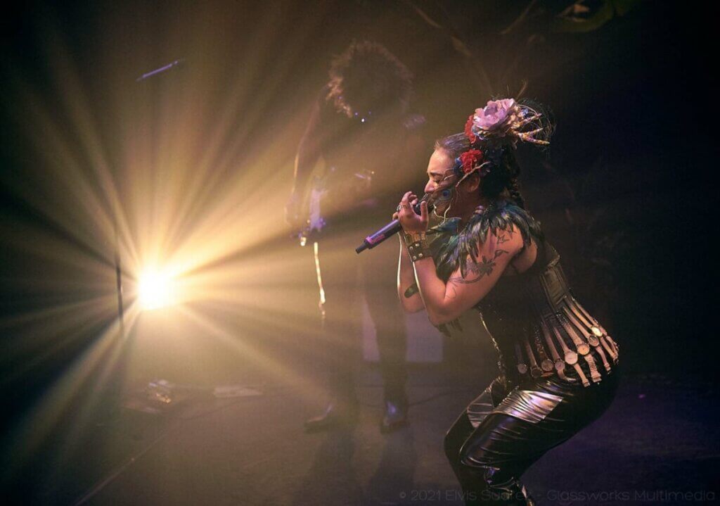 Sol "La Barbara" wears flowers and a peacock feather in her hair as she sings passionately into a microphone. She is also wearing a feathered collar and a corset with tan watches around the waist. In the background Rey plays "Guarapo" on the guitar with his head down. There is a spotlight on them both.