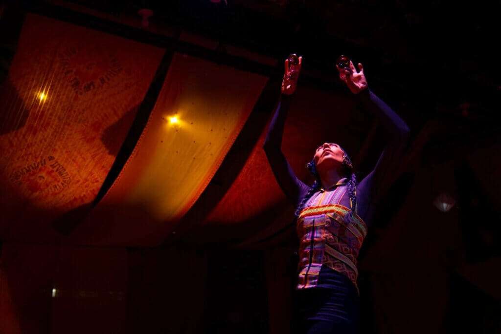Nejla Yatkin - Ouroboros A dancer standing and reaching arms upwards in colorful top with colorful fabric in the back hanging from the ceiling.