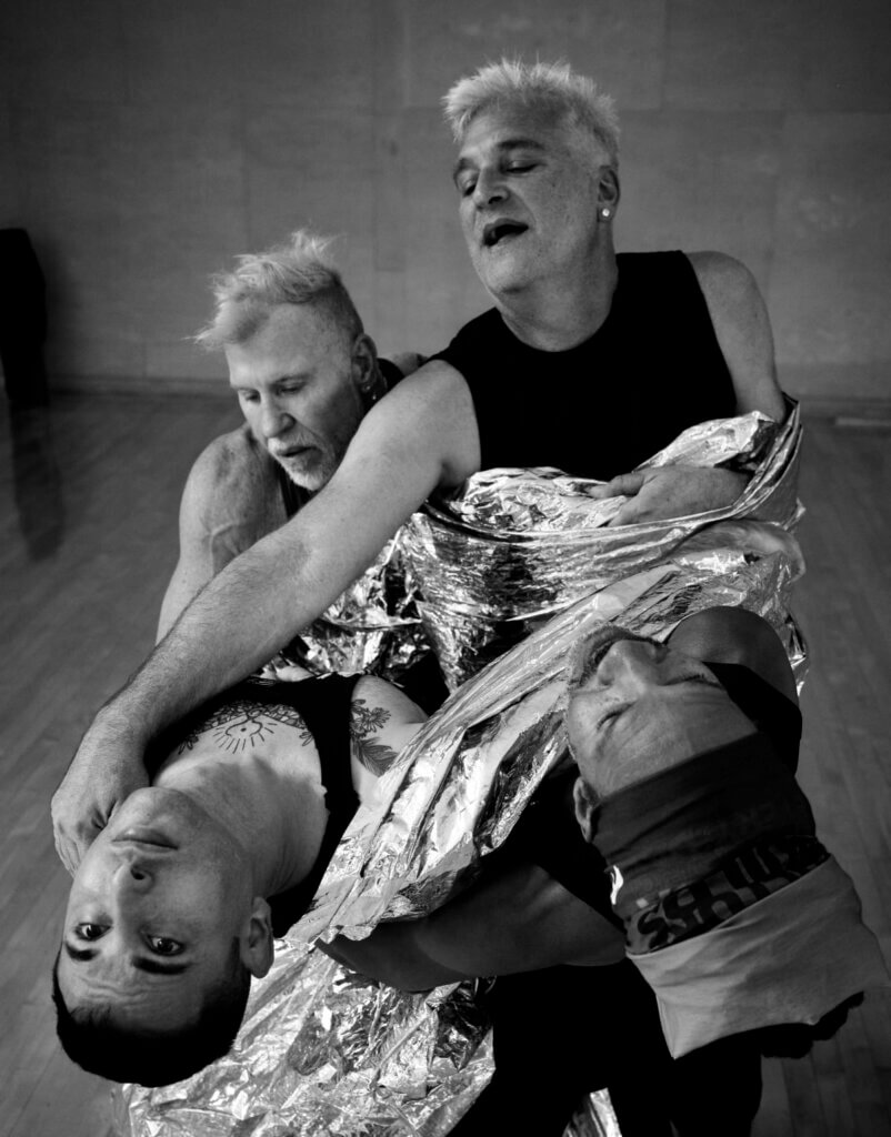 Pioneer Winter - Apollo Four dancers in dramatic counter-balance, connected by reflective fabric: Pioneer Winter, a 36-year-old white genderqueer with chest tattoos, arches back alongside Clarence Brooks, a 64-year-old Black genderqueer with dreadlocks. They are supported by Cuban-American 59-year-old Octavio Campos with silver hair, and 66-year-old white Frank Campisano, also with short silver hair.