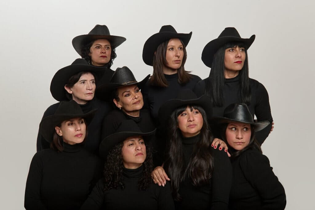 Dora Arreola & Mujeres en Ritual Danza-Teatro - Queering the Border Nine Mexican women dressed in black, wearing black cowboy hats, huddle together and lean on each other, gazing off camera to the right, with serious expressions, and serious attitude.