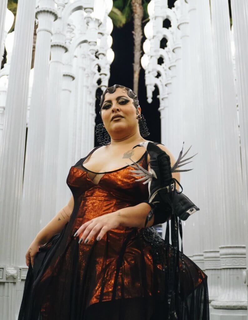 San Cha LACMA A medium-skinned Latinx performer stands in front of white columns. They wear a dark amber dress, fitted with a corset. On the performer’s left arm, they showcase a bird puppet with silver feathers. Their hair is in an updo with waves, gold and black eye makeup, and large black earrings.