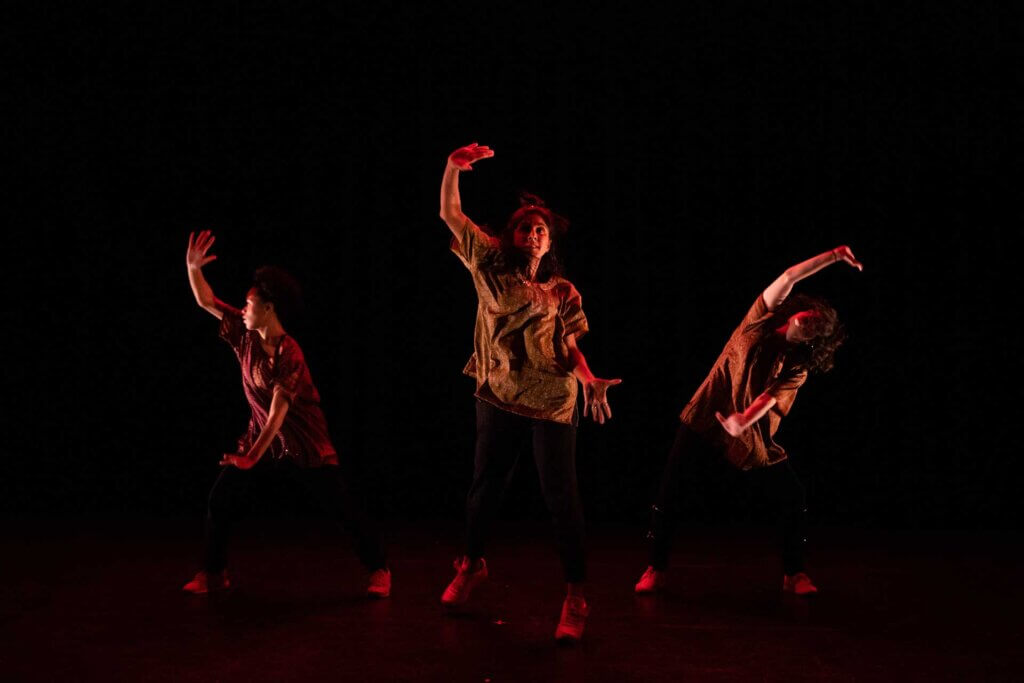 Three women are posed in this image with their right arms up high and left arms stretched low. The women on the left and right sides are side bending away from each other, while the woman in the center is facing forward. The woman on the left is a medium-skinned Black woman. The woman in the middle is a medium-skinned Indian woman, and the woman on the right is white. The women are wearing similar shirts and darker bottoms and the background is black.