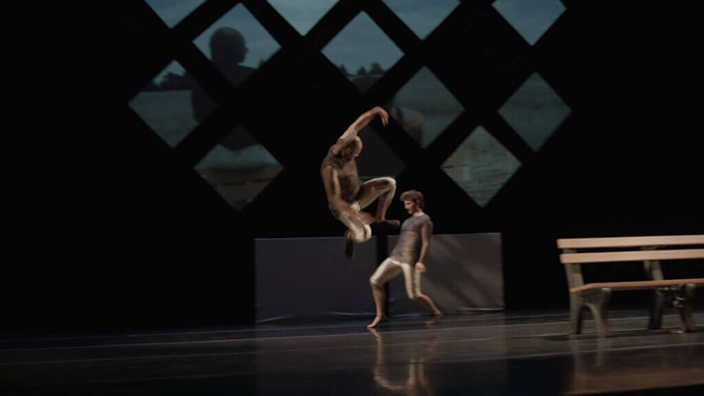 An interracial male duet (one white male, one Black, medium light-skinned male) in blue, partially see-through knit tops and gold football pants on stage with a set that includes a park-style bench and diamond and rectangular shaped projection surfaces upstage. The Black male dancer is downstage in a jump with legs bent and left arm extended above, and white male dancer is upstage in profile leaning back.