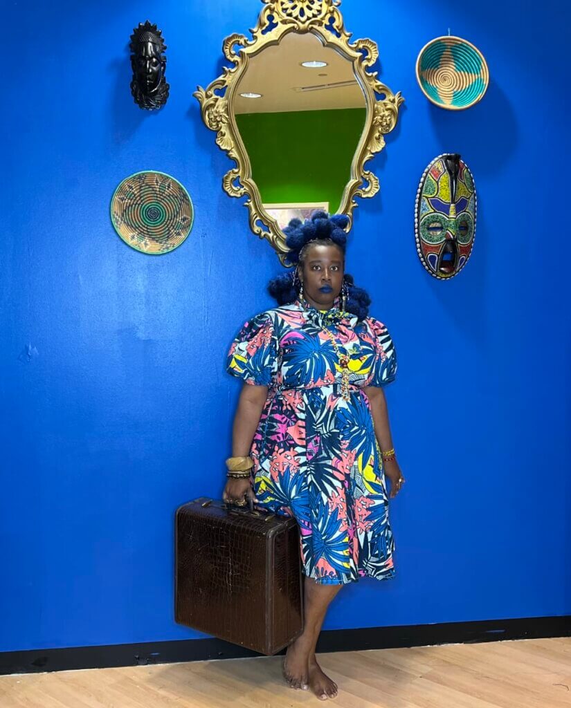Ifátùmínínú Bámigbálà Arẹ̀sà This image shows a dark-skinned, barefoot woman with blue hair styled in tribal braids. She is wearing a blue Ankara dress against an ultramarine blue wall and holding a vintage letter suitcase. African masks, a large mirror, and baskets hang on the wall.