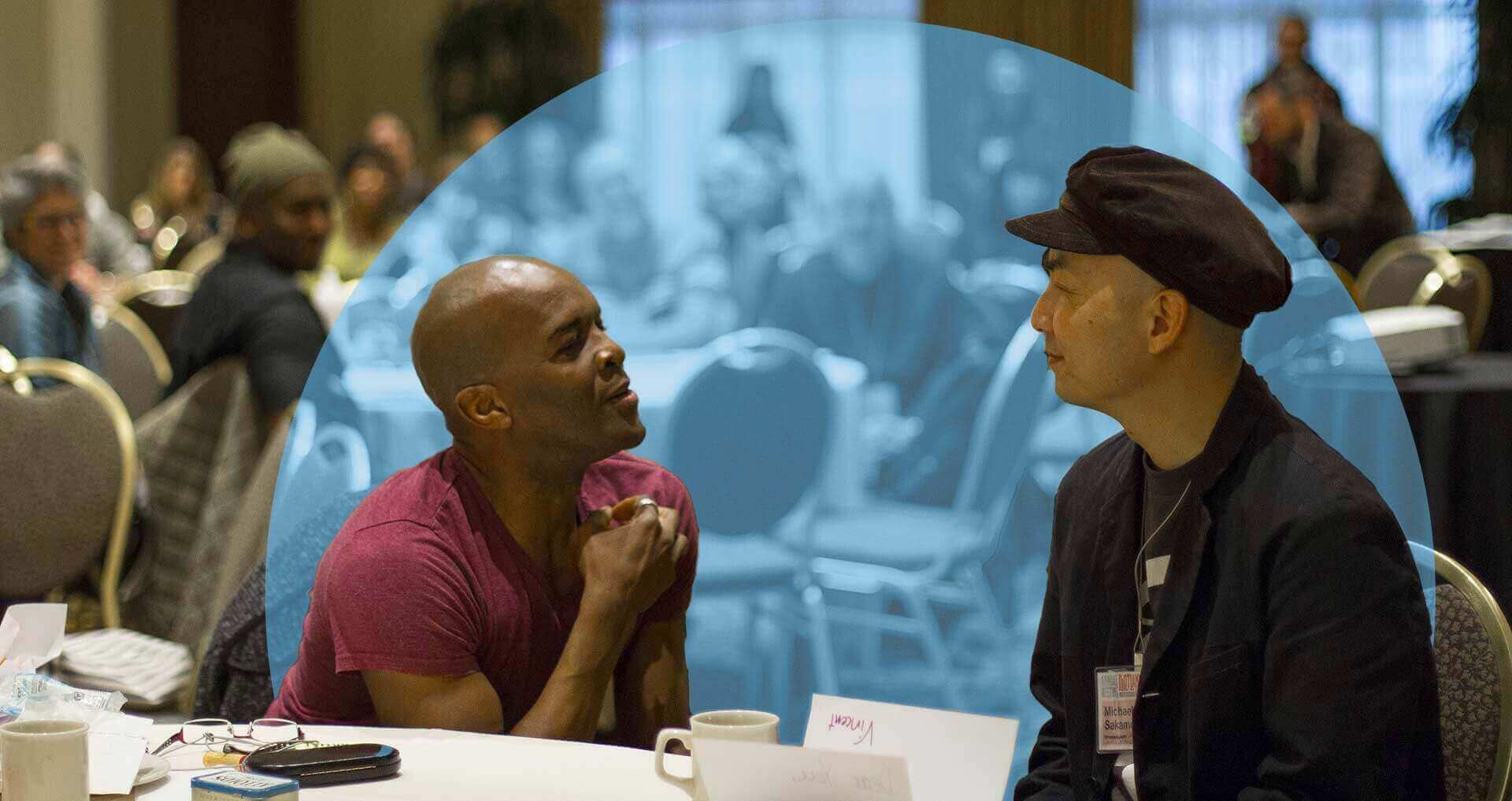 Two men sit face-to-face at a round table in a conference setting, engaged in an attentive and animated conversation. The background shows other attendees blurred and tinted blue within a digitally added semicircle shape. One man wears a red shirt and gestures with his hand, while the other, dressed in black with a cap, listens intently.