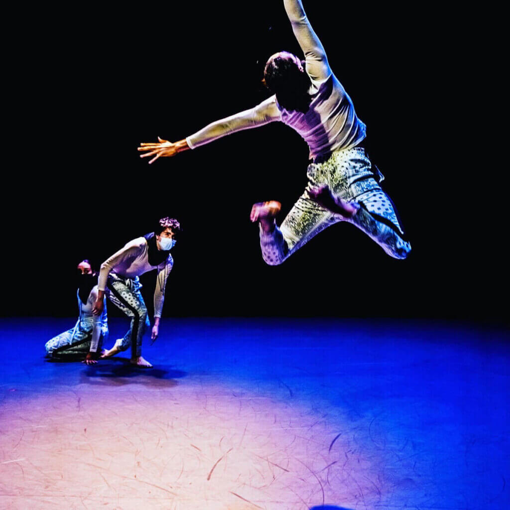A medium-light skinned Black woman is sitting next to a crouching fair-skinned white man who is ready to run toward a tall medium-light skinned Black men. The tall Black man with is back to the viewers is leaping into the air, his knees are bent, right arm extending to the sky, left arm extending toward the Black woman and white man. They all are wearing white long sleeves shirts, white joggers pants with a bandana print, ski masks around their necks, and NK95 masks.