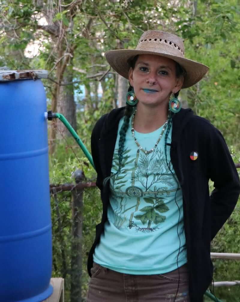Jenna Fricke This image is of a light-skinned/tan femme person wearing work pants, a light green shirt, and a straw hat standing outside next to a rain barrel connected to a house's gutter system. They styled their/her dark hair in two long braids, and they are wearing green Indigenous beadwork earrings and bright blue lipstick. Trees are in the background.
