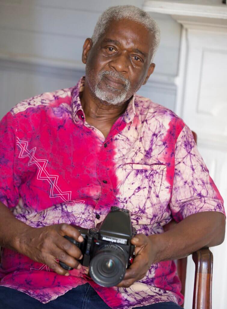 Keith Calhoun Photo - Photogragher Chandra McCornick This image shows a dark-skinned African American man with gray hair and a goatee. He is sitting in a wooden chair, holding his camera while looking at the viewer. He is wearing a tie-dyed fuchsia, purple and white button-down shirt and dark blue jeans. The background is a gray wall and a white-painted fireplace mantle.