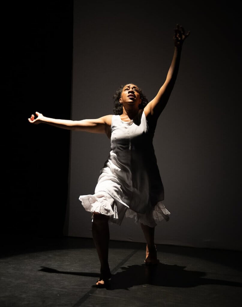 Kesha McKey The image of a light-skinned African American woman wearing a loose white knee-length tank dress in mid-movement while dancing under a white spotlight on a black box stage. She is in a lunge position, with her arms up and side. Her gaze is upward, and her hair is in relaxed curls.