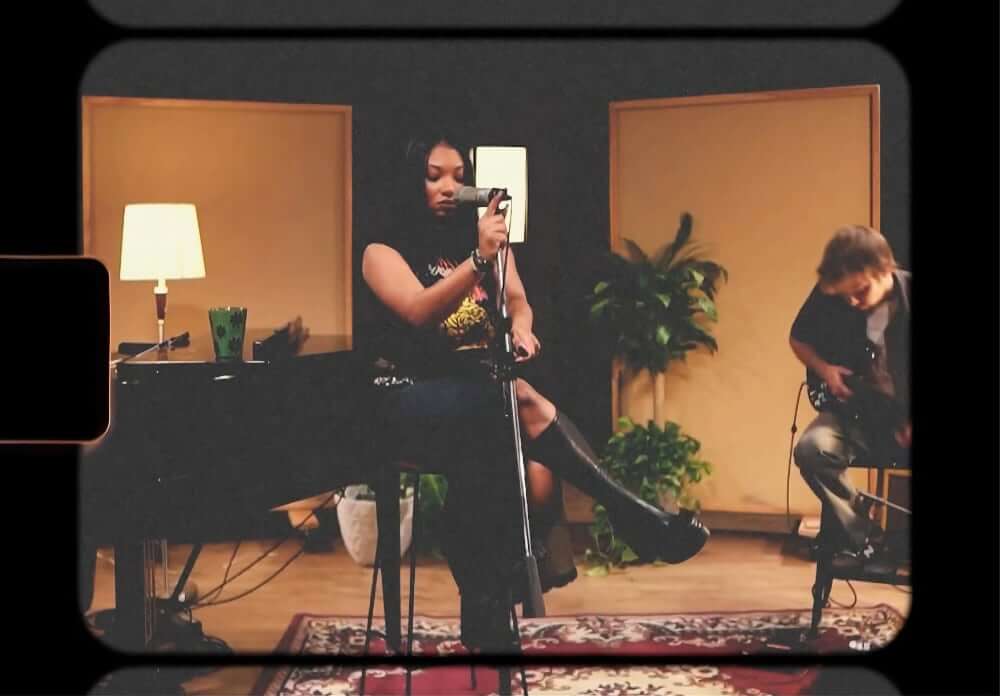 Kiana Tenille - Homesick Live The image of a medium-skinned American American musician sitting cross-legged on a stool while holding a microphone in a dimly lit recording studio. To her left sits a pianist, and to her right is a guitarist. Lamps and plants are in the background, and a cup of tea is atop a black baby grand piano.