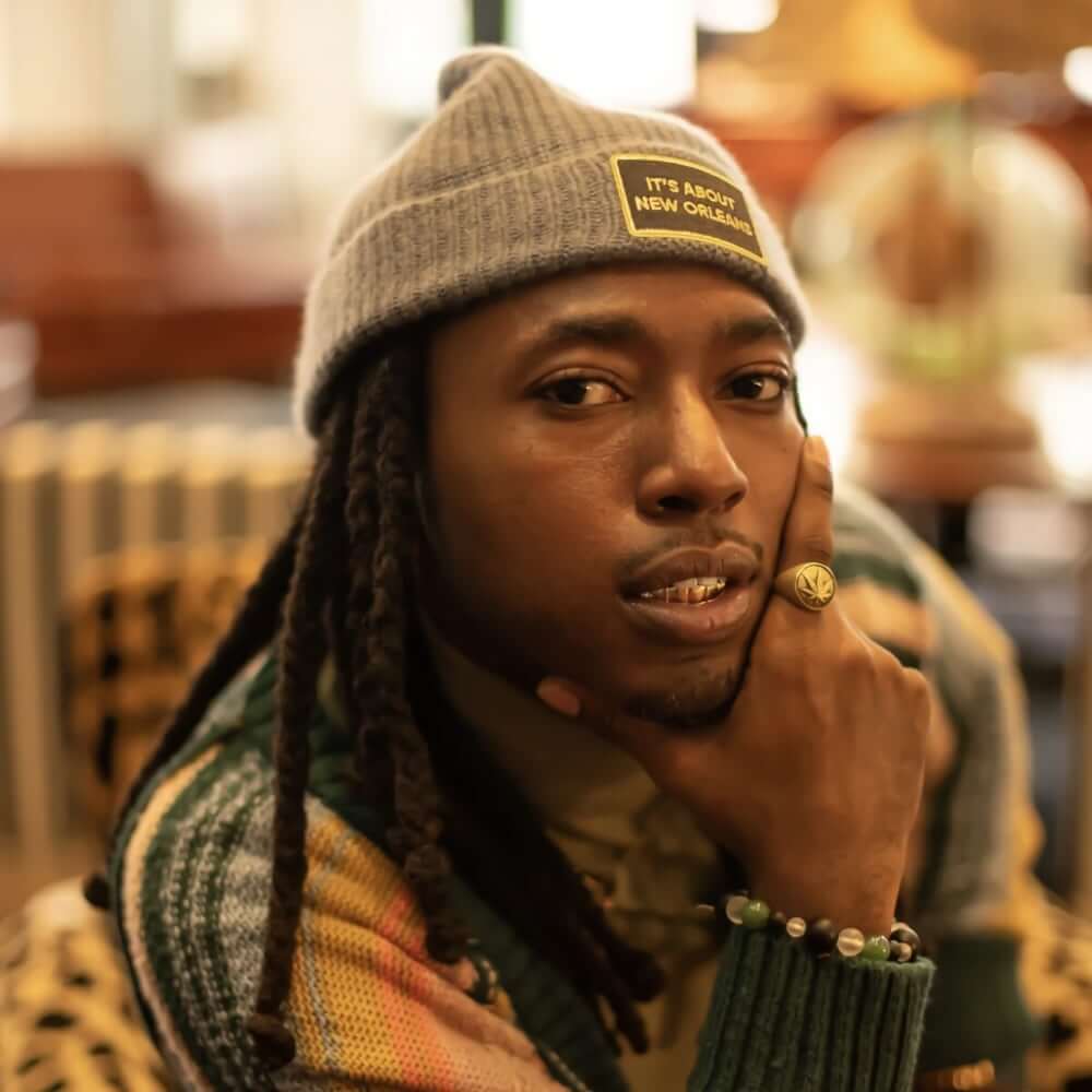 Kr3wcial An image of a medium-dark-skinned African American male with a goatee and long locs. He wears a grey knit cap and a multi-colored striped sweater. He looks into the camera, his chin resting in his right hand.