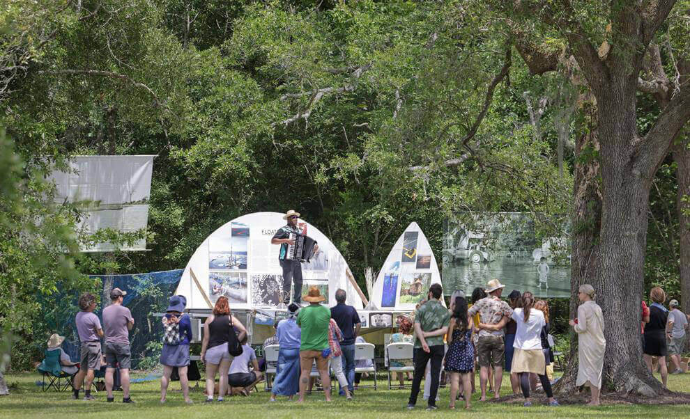 A medium-skinned accordion player with a black short-sleeved shirt and a straw hat is standing on a partially constructed boat called the Float Lab. He is singing while looking out at a crowd of about 50 people.