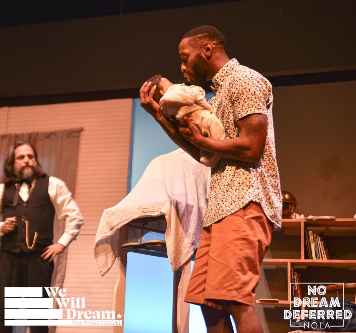 In a living room, A young Black man wearing a light blue button-up shirt and golden orange shorts, holds his small baby, while the ghost of abolitionist JOHN BROWN, portrayed by a White man, with a full beard and shoulder-length hair, wearing a black vest, white collared shirt, and black pants, visits behind him, in the wonder of the small child.