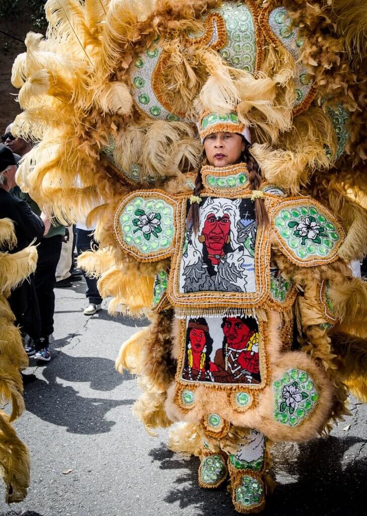 Laurita “Big Queen Rita” Dollis The image is a full-body portrait of a light-skinned African American woman wearing a gold-feathered Black Indian suit. Her headdress has gold feathers, green and white beads, and large green rhinestones. The stonework patches on her arms are of green and white magnolias, and the front patches show human figures in red, brown, white, and black beads. She is standing on the street surrounded by a crowd of adorning fans on a sunny day.