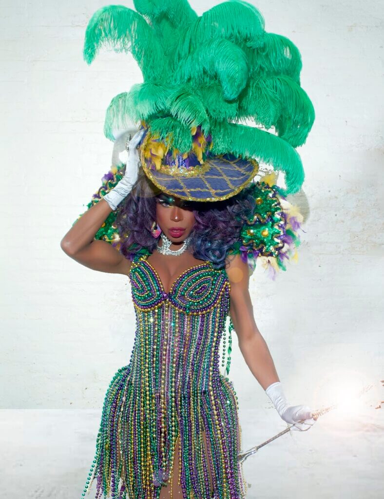 Laveau Miller An image of a medium-skinned African American woman wearing an extravagant Mardi Gras-inspired outfit. The ensemble features a beaded corset dress adorned with purple, green, and gold beads, forming a fringe-like pattern, white opera gloves, and a purple hat with gold trim and large ostrich feathers. The background is a plain white wall, and she holds a baton in one hand.