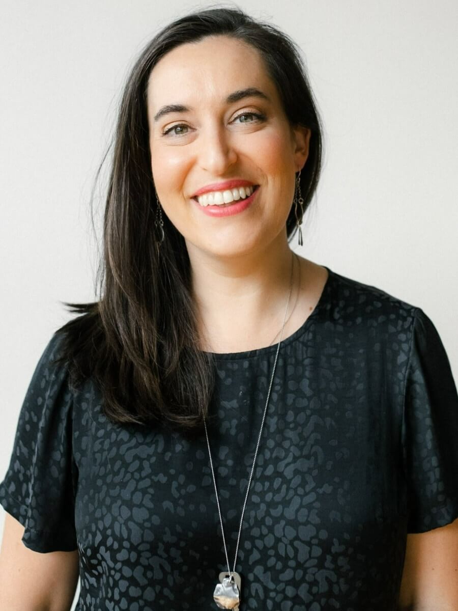 The photo shows Katie Collins, a light-skinned woman with black shoulder-length hair, smiling and directly facing the viewer, for a headshot photo. She wears a black shirt with subtle designs, along with a long, silver necklace and dangling earrings.