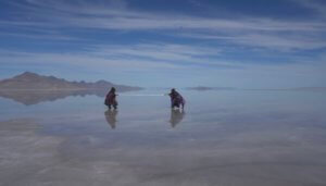 Two figures in the middle-distance in a desert landscape, with mountains in the far background and water on the ground reflecting a bright blue sky. They are dressed in Afro-futurist costumes and each pull on a white rope, as in the child’s game “tug of war.”