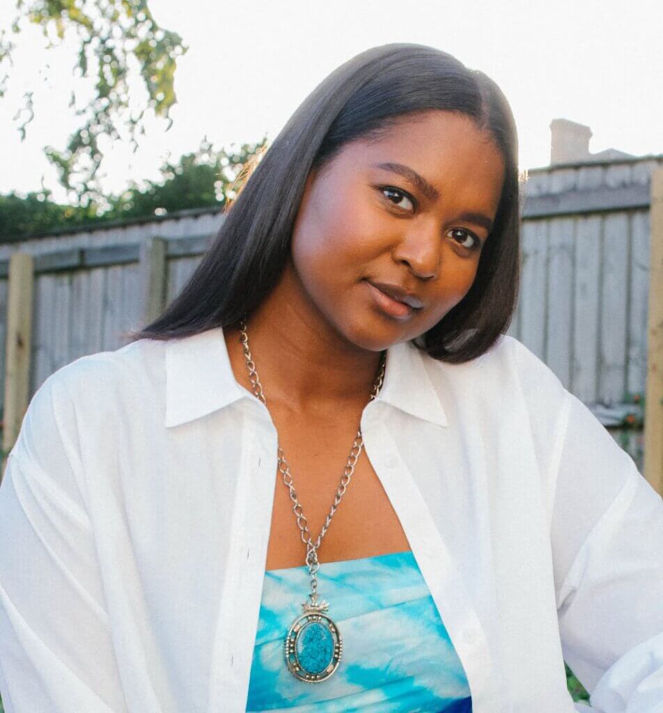 Maaliyah Papillion The image shows an Indigenous woman with long, black, straight hair. She is looking at the camera with her head tilted to the side. She wears a white button-down shirt, an aqua tank underneath, and a silver and turquoise jewelry necklace.
