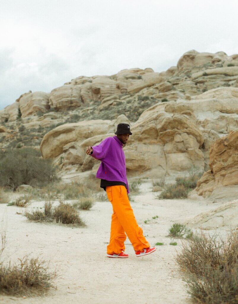 Malik Sanders This image shows a dark-skinned man in mid-motion in the desert. His right arm is raised to shoulder level, and his right foot is slightly raised off the ground. He wears orange pants, a purple sweatshirt, and a black hat.
