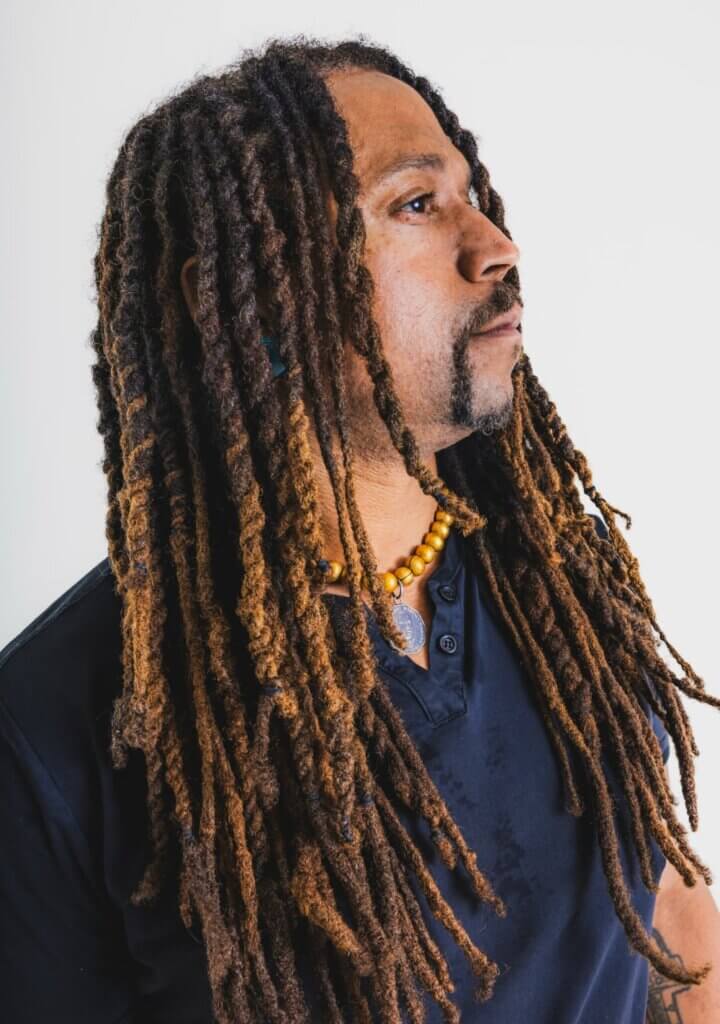 Marcus Brown A light brown-skinned African American man standing with his head tilted to the sky. He has long, brown, twisted locs and a gold-beaded necklace around his neck.