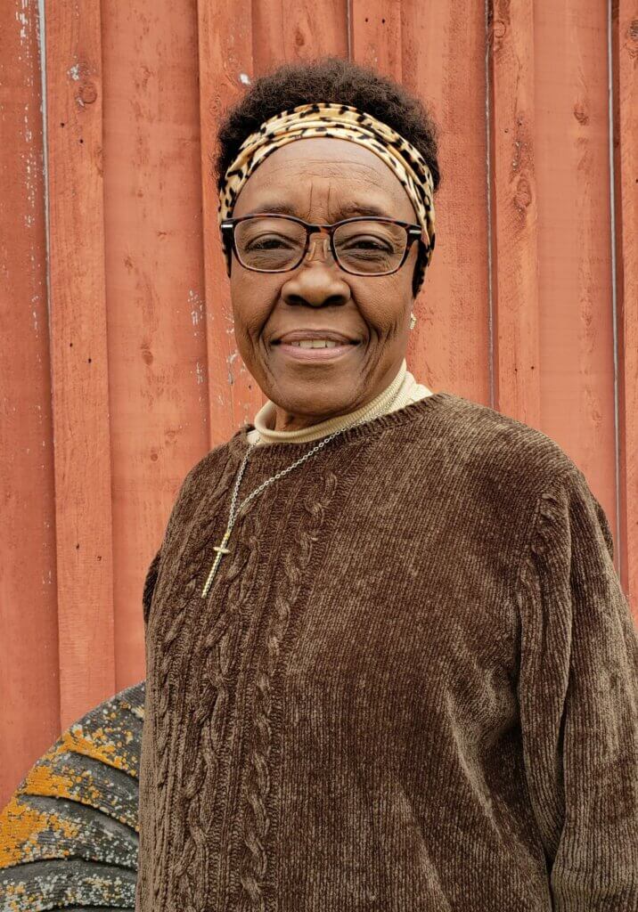 Mary Victorian Jackson The image is of a medium brown-skinned African American woman. She is wearing a brown velour sweater, tan turtleneck underneath, black-rimmed eyeglasses, and a leopard print headband. The woman is standing outside in front of an old red barn.