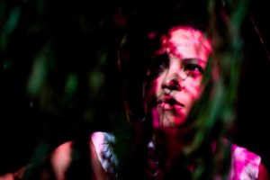 A medium-light skinned woman with dark eyes and hair wearing a white top looks upward through green leaves, shadows, and dark pink light. Her face is in focus and partly in shadows, while leaves around her face are out of focus. The background is black.