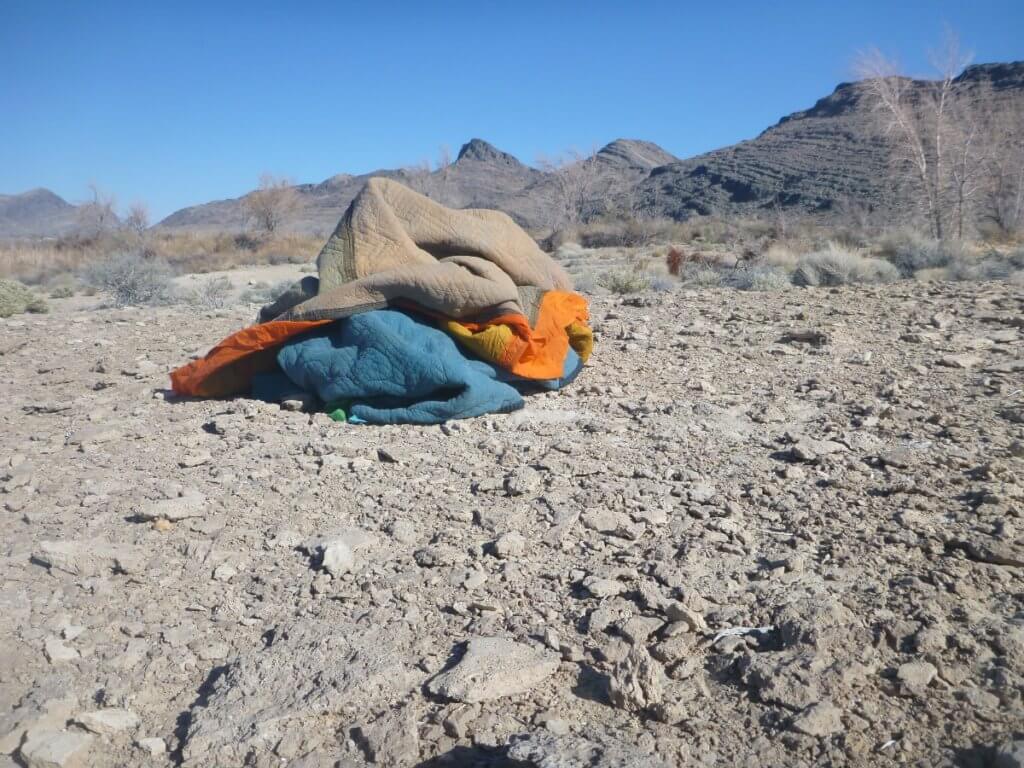 A pile of blankets and fabric in orange and blue is situated on a rocky hill in front of a mountain range. The peak of the blanket pile is shaped to match the peaks in the mountain range and the patterns on the blanket echo the lines of the mountains and rocks beyond.