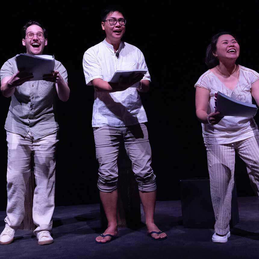 A trio of actors onstage with scripts in hand: a white man with glasses, a medium light-skinned Filipino man with glasses, and a petite medium-skinned Filipino woman stare up to the sky, wide, joyful smiles on their faces. They wear pants and shirts in whites, tans, and light browns.
