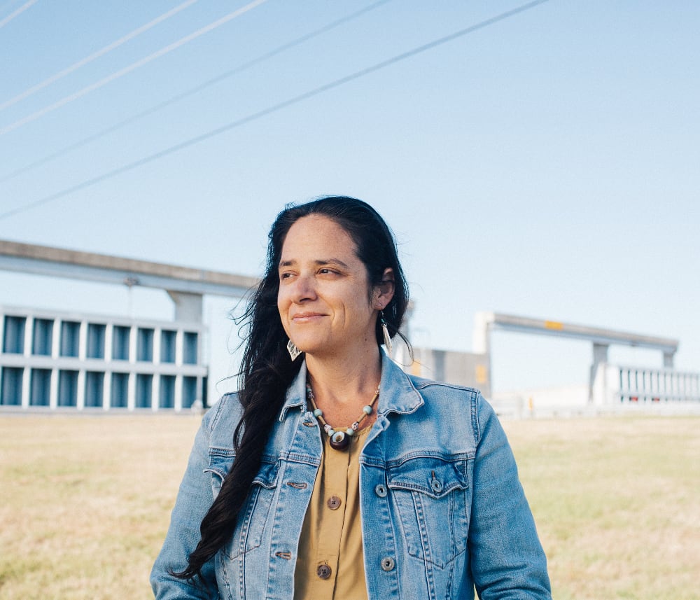 Monique Verdin An image of an Indigenous woman in a jean jacket standing before a concrete flood levee wall. She has long brown hair, a mustard-colored blouse, and a seed pod necklace.