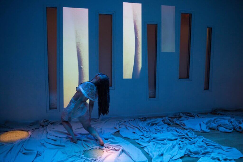 A light brown-skinned Black woman leans back, her midback length locs dangling in the space behind her. She is wearing a rumpled, paper-thin plastic set. Large scattered and scrunched panels of canvas are beneath her and projected images of a fold or crevice in a body of similar complexion are behind her.