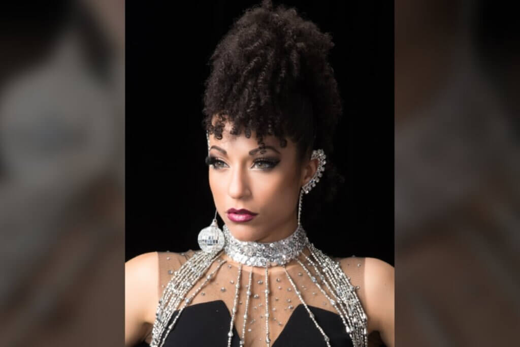 An olive-skinned, multiracial woman with dark hair and hazel eyes is shown from the shoulders up. She has tight curls pulled up high on her head with bangs and is wearing a black strapless top with rhinestones lining her neck area. She has a silver, sequined choker that drips several strips of silver beads past her top. She wears a dangling, disco ball earring in one ear and the other ear is lined with silver rhinestones that cover the entire ear and drip down into a single strand from the earlobe.