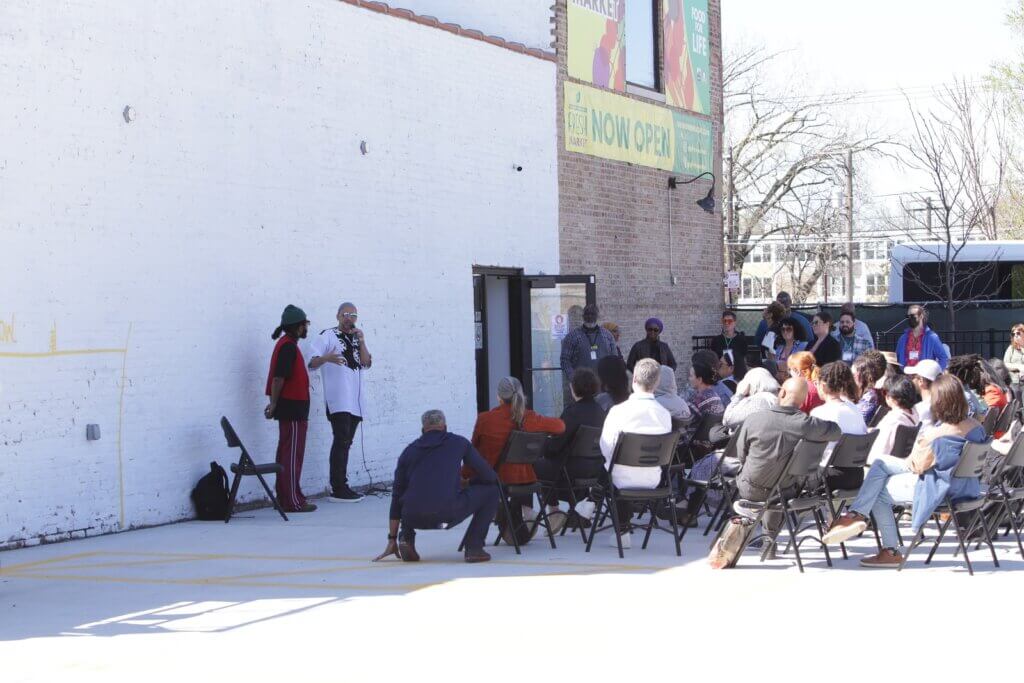 Two men stand in shadow against a white painted brick wall of a building; one of them is speaking into a microphone. Before them, a group of people sit in rows of folding chairs and listen.