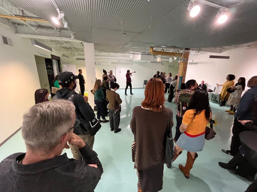 A group of people standing in random positions throughout a brightly lit, large white room with a low ceiling and no windows. The view is from the back of the group so that most of the people in the photo are shown from behind. They all appear to be listening to a person at the other end of the room who is speaking into a microphone while gesturing to something off to the side.