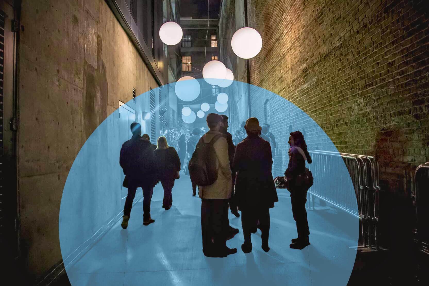 A group of people stands and walks through a dimly lit urban alleyway decorated with glowing white globe lights suspended overhead. A soft blue circular overlay highlights the center of the image, drawing focus to the figures in the middle of the walkway. The alley is framed by tall brick and concrete walls, giving the scene a cozy, urban-night feel.
