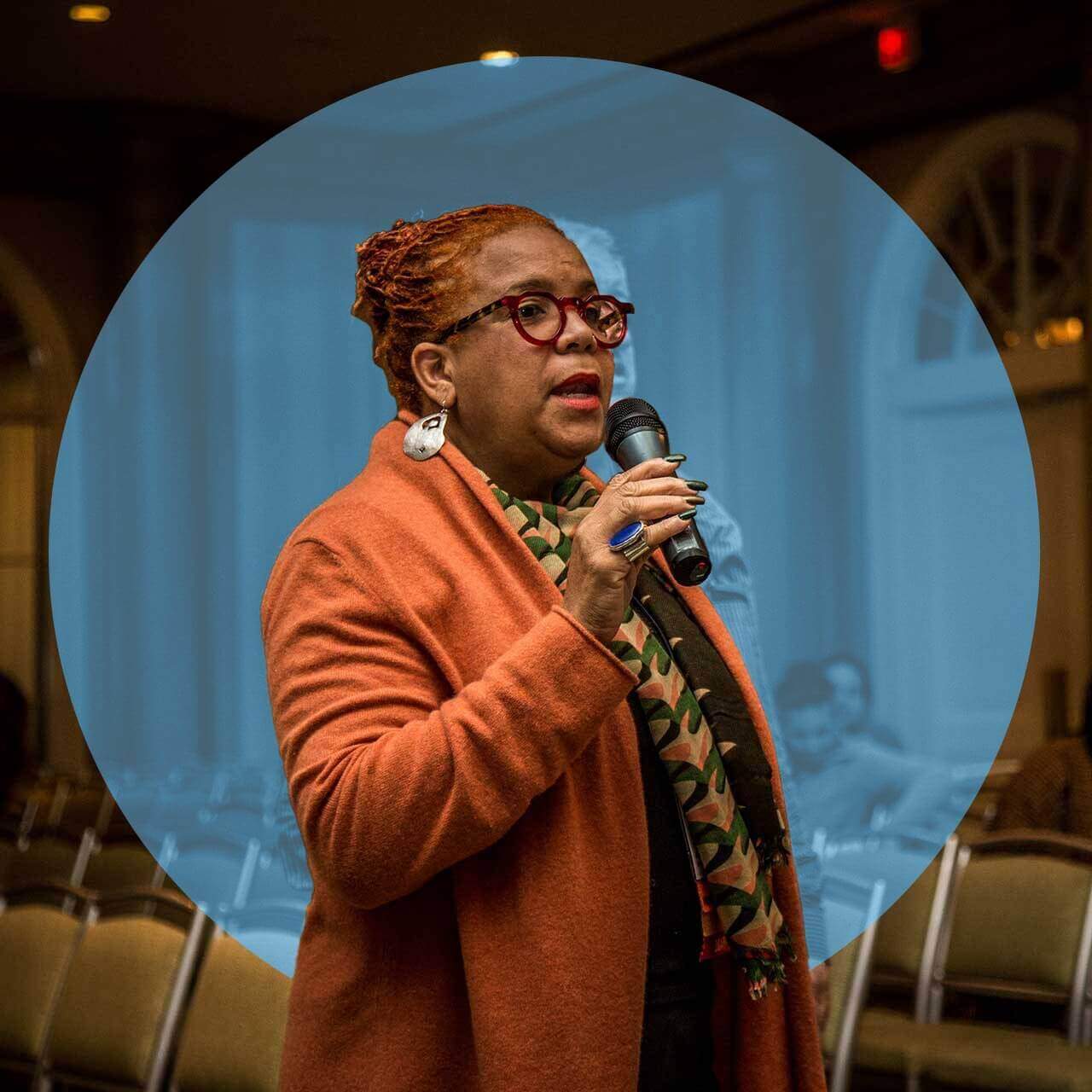 Rosie Gordan-Wallace, a brown-skinned woman wearing an orange coat and patterned scarf, speaks into a microphone during an event, standing among rows of empty chairs. She is framed by a blue circular overlay that emphasizes her presence in the image. Her expression is focused and engaged, suggesting she is participating in a discussion or giving a presentation.