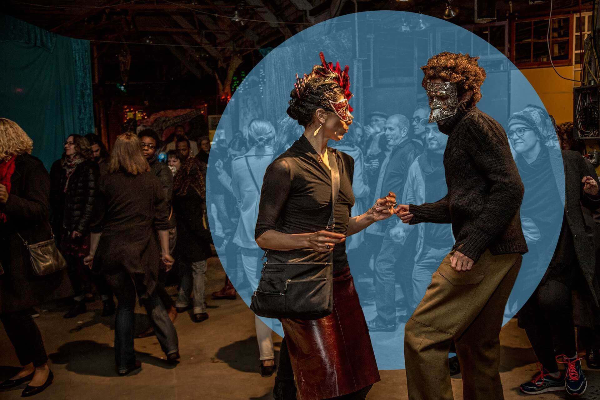 Two people wearing elaborate masquerade masks dance together in the center of a dimly lit event space, highlighted by a blue circular overlay. The background reveals a crowd of attendees mingling and dancing, with moody lighting and eclectic decor setting a festive, theatrical vibe. The couple’s expressive body language and costumes suggest a creative, celebratory gathering.