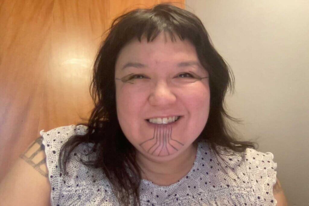 A woman with tribal tattoos is smiling with all of her heart and love against a brown and grey background. She is wearing a black and white dot-printed shirt and one of her traditional shoulder tattoos peeks through on one side. She has black hair, black eyes and is slightly overweight.