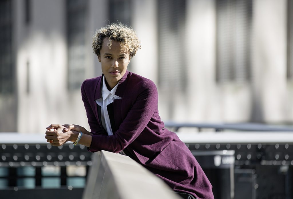 Kaneza Schaal poses leaning on a bridge in a white shirt and purple sweater