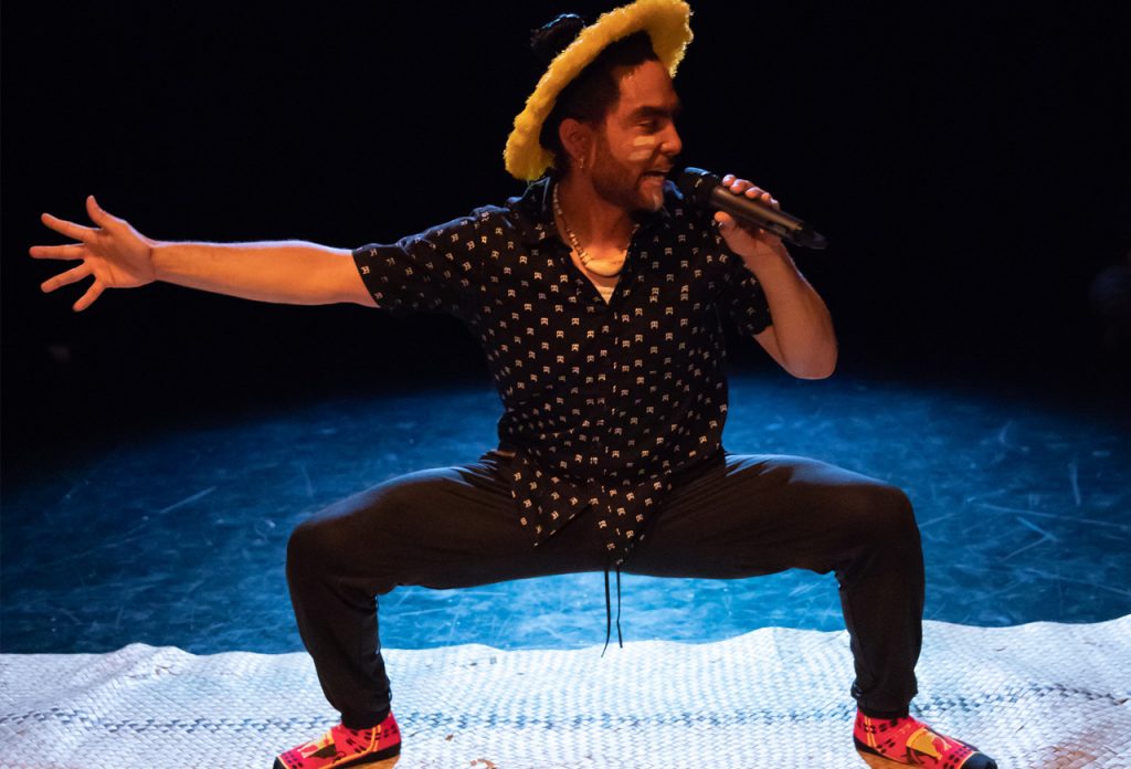 Dakota Camacho squats, face turned to a microphone, right arm extended. Camacho wears black clothing, facepaint, and a yellow and black hat.