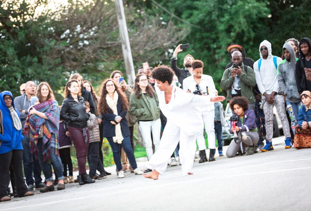 Onye Ozuzu, wearing white flowing clothes, dances outdoors in front of a crowd